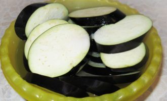 Как быстро и вкусно запечь баклажаны в духовке? Для начала хорошенько промойте свежий баклажан и нарежьте его на кружочки толщиной примерно по сантиметру. Кружочки опустите в объемную миску и залейте очищенной подсоленной водой на полчаса, чтобы сошла горечь.